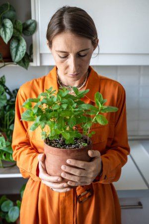 Scandinavian florist woman in orange dress holding flower pot with fresh green mint grown at home. Gardener amateur plant lover growing edible fresh herbs. House planting, hobby, eco gardening.