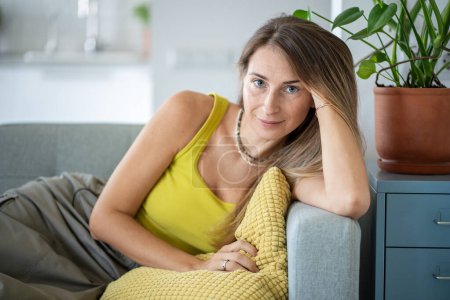 Serene carefree woman having rest after work looking at camera, leaning back on back of sofa at home. Portrait relaxed female spending lazy time at weekend in living room. Mindfulness and slow life.