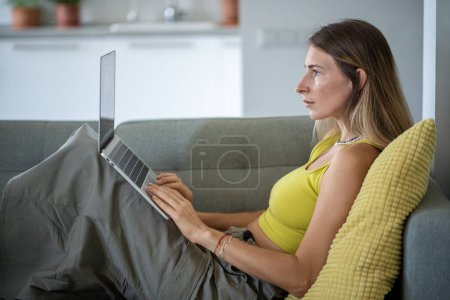 Photo for Focused freelancer woman online working on laptop, lying on sofa at home. Concentrated female in yellow top using computer on couch, at living room. - Royalty Free Image