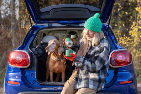 Photo for Woman pet owner pouring tea from thermos sitting in car trunk with vizsla dog on nature in forest in road trip. Autumn outdoor activity, Scandinavian vacation, travel tourism weekend with beloved dog. - Royalty Free Image