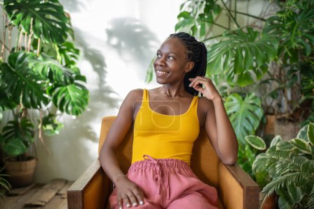 Téléchargez les photos : Heureuse jeune femme noire se détendre sur une chaise dans un confortable appartement vert avec des plantes de monstère, joyeuse fille afro-américaine passer du temps, regardant de côté, se reposant dans un fauteuil au jardin à la maison. Sans stress - en image libre de droit