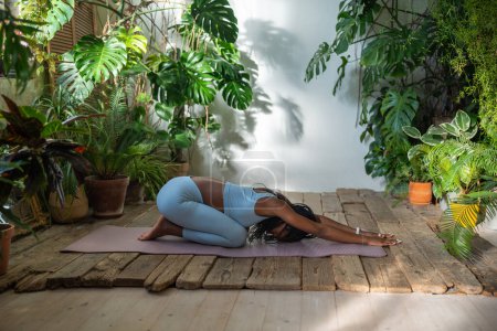 Téléchargez les photos : Axée sur la respiration, la femme afro-américaine allongée sur un tapis de fitness s'étirant vers l'arrière, relaxant les muscles après l'entraînement à la maison. Athlétique femelle noire développe la flexibilité du corps pratiquant dans un studio de yoga confortable. - en image libre de droit