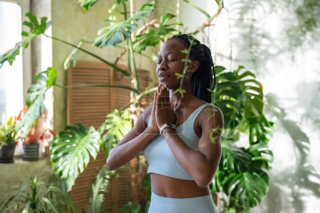 Téléchargez les photos : Femme noire concentrée méditant dans le stand de pose de yoga avec les yeux fermés dans le jardin à la maison avec Monstera, trouver la tranquillité et le calme intérieur. Africaine fille pratiquant vrikshasana et exercices de respiration. - en image libre de droit