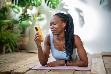 Téléchargez les photos : Satisfait femme afro-américaine détendue tenant smartphone couché sur tapis de fitness yoga se reposant après l'entraînement. Femme noire athlétique positive regardant l'écran téléphone portable prendre une pause dans l'entraînement sportif - en image libre de droit