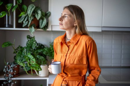 Foto de Mujer reflexiva sosteniendo taza de pie en la cocina rodeada de plantas de interior. Retrato de mujer tranquila y confiada en vestido naranja sostiene la taza de té, pensando, mirando a la ventana. - Imagen libre de derechos