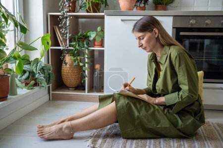 Konzentrierte Frau aufmerksam schreibt persönliches Tagebuch sitzt auf dem Küchenboden zu Hause. Nachdenkliches, ernsthaftes, weibliches Denken, das Text im Notizblock erschafft. Ideen entwickeln, sich selbst beobachten, Gedanken aufschreiben