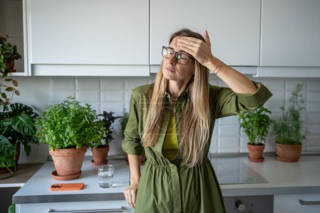 Photo for Tired exhausted blonde woman feels headache touching head with closed eyes on kitchen at home. Unhealthy female in glasses suffers head pain, dizziness, migraine, cephalalgia feeling unwell pressure. - Royalty Free Image