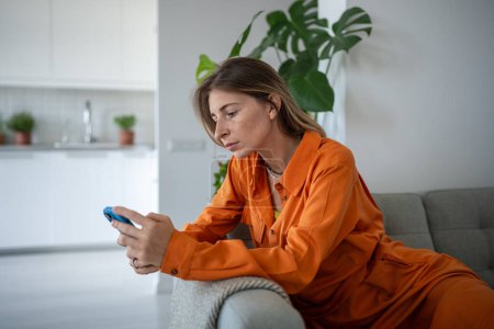 Photo for Lonely fatigued woman sitting on sofa holding smartphone sadly looking at screen scrolling web social media. Tired middle aged scandinavian female waiting message or call feeling upset, frustration. - Royalty Free Image