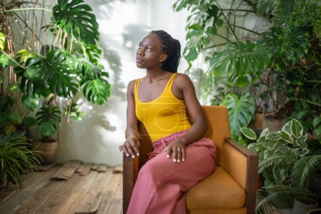 Foto de Calma joven mujer negra relajarse en la silla en acogedor apartamento verde con plantas monstera, niña afroamericana reflexiva pasar tiempo, pensando mirando a un lado, descansando en el sillón en el jardín en casa. - Imagen libre de derechos