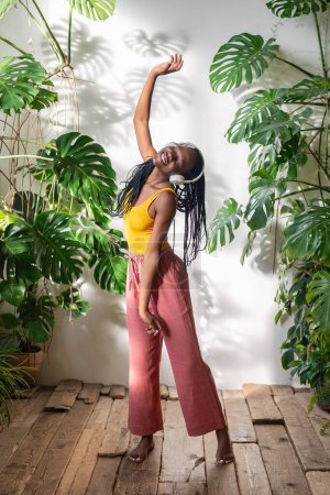 Carefree black woman dancing barefoot in headphones listening to music on wooden floor with pleasure, enjoy moment. Relaxed african american young female chilling out to songs in room with houseplants