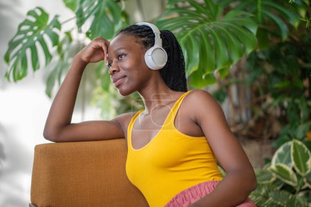 Relaxed african american woman resting in house garden use wireless headphones listening music enjoy weekend surround by houseplants. Dreaming black young female rest to reduce stress in greenery room