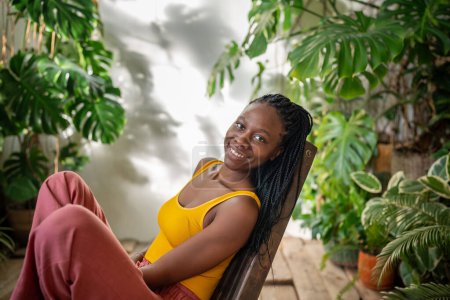 Photo for Contented carefree african american woman sits on chair, surrounded by tropical houseplants as looks at camera. Serene relaxed black girl smiling reclines in comfortable armchair in urban jungle room. - Royalty Free Image