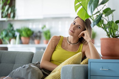 Portrait of woman on cozy sofa among plants at home. Calm female resting relaxing at couch, looking at camera with confident sure quiet serene face expression, leaning on armrest, enjoying comfort.