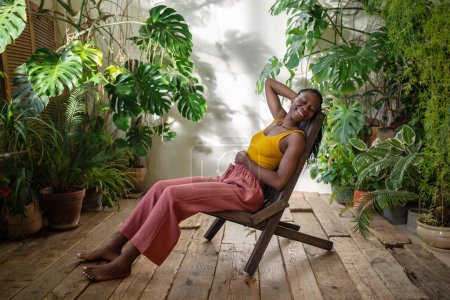Foto de Mujer afroamericana alegre con sonrisa que refleja el disfrute de descanso muy necesario se sienta en la silla en el ambiente soleado de la sala de la selva urbana. Agradable hembra negra descansando en el salón de casa con plantas de interior. - Imagen libre de derechos