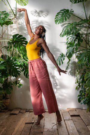 Serene African American young woman relaxing to songs amidst lush houseplants, savour moment. Carefree black woman dancing barefoot in headphones, immersed in music on wooden floor experience pure joy