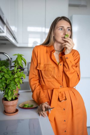 Photo for Woman sniffs home-grown basil leaf. Female gardener enjoying smell of green basil plant. Girl in the orange dress grows spicy herbs at home for cooking. - Royalty Free Image