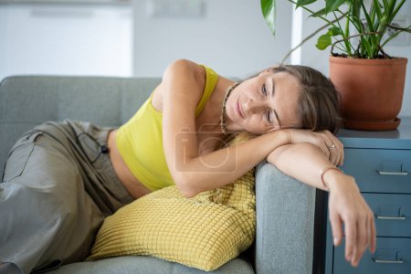 Müde Skandinavierinnen liegen tagträumend auf der Couch zu Hause. Erschöpfte Frau entspannt auf der Armlehne des Sofas liegend und über die Zukunft nachdenkend. Psychische Gesundheit, Stressabbau, Verzögerung.