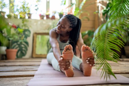 Téléchargez les photos : Sport pour un corps sain. Athlétique femme noire développer la flexibilité assis sur tapis de yoga entourer de plantes tropicales d'intérieur. Plaisir afro-américaine fille tenant les pieds avec les mains faisant des exercices de remise en forme. - en image libre de droit