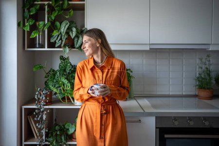 Photo for Smiling carefree woman drinking tea in kitchen. Happy satisfied delighted girl among houseplants on the kitchen enjoying morning time, looking out window, holding mug of coffee. Slow life concept - Royalty Free Image