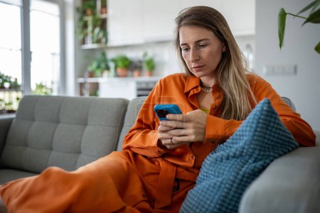Relaxed woman use cellphone sit on sofa order delivery at home. Focused female look at smartphone screen make online purchases, scrolling web, surfing social media, internet chatting resting on couch.