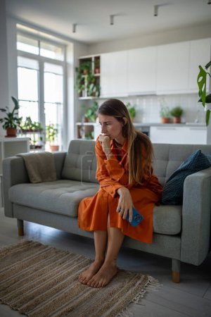 Photo for Stressed sad woman sits on couch reflecting deep in thought. Serious, melancholic, focused female with cellphone, put hand to chin, looks at floor, thinking mentally solves problems, pondering at home - Royalty Free Image