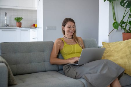 Pleased woman smiling using laptop on knees sitting at couch. Interested female with computer on lap have fun online enjoying comfort, chatting with friends on social media networks, watching video.