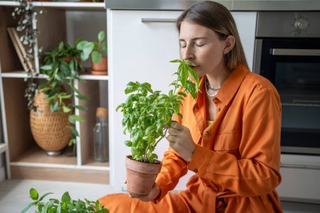 Photo for Woman enjoying smell of fresh basil. Girl in orange dress sits on floor, inhales aroma of home-grown flavour basil in pot. Female gardener sniffs aroma herbs, closed eyes - Royalty Free Image
