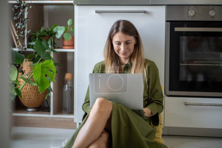 Photo for Satisfied woman using laptop on knees sitting on kitchen floor. Smiling happy female looking screen at portable computer, taking online course, watch webinar, listen to internet lecture, remote works. - Royalty Free Image