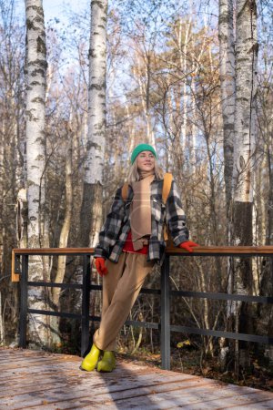 Foto de Sonriente senderista serena de mediana edad que descansa en el parque de otoño disfrutando de la naturaleza escandinava. Despreocupada mujer caminando en el bosque de pie en el puente disfrutar de la naturaleza. - Imagen libre de derechos