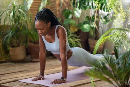 Active african american woman stretching with closed eyes on fitness mat surround by tropical indoor plants. Sported black female do yoga, develop flexibility for healthy body in cozy sported studio.