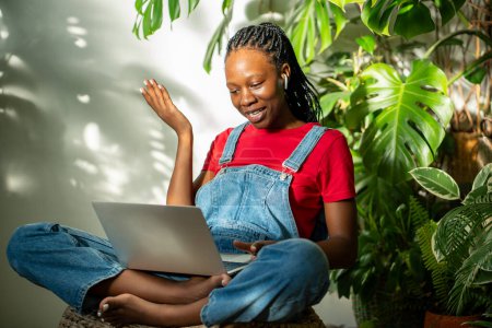 Photo for Content African American young woman with laptop resting on knees and earphones, participates in video conversation call with friend, records a new vlog, gesturing with hand at home with houseplants. - Royalty Free Image