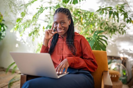 Foto de Mujer afroamericana alegre sonriente mirando a la cámara con el ordenador portátil en las rodillas sentado en el sillón rodeado de plantas de interior. Agradable mujer negra freelance trabajando en un acogedor espacio de coworking con computadora. - Imagen libre de derechos