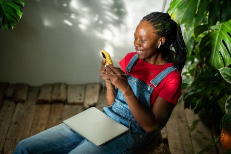 Glückliche afrikanisch-amerikanische Frau mit Kopfhörer-Look Screen-Smartphone hören Podcast sitzen mit geschlossenen Laptop auf Knien umgeben von tropischen Pflanzen. Gefallen schwarze Mädchen nehmen Pause mit Musik auf dem Handy.