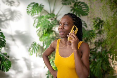 Foto de Pensativo mujer afroamericana interesada hacer llamada con teléfono móvil rodeada de plantas tropicales verdes de interior mirar a un lado ventana. Centrado en la conversación chica negro hablar en el teléfono celular con el novio - Imagen libre de derechos