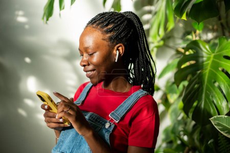Photo for Contented african american woman wear earphones, looks at smartphone screen, engrossed in selects favourite music track on streaming app. Black girl holds phone scrolling social media at home garden. - Royalty Free Image