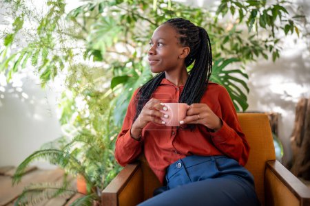 Relajado soñando mujer afroamericana sosteniendo taza de café sentado en la silla en la selva urbana interior mirando a un lado de la ventana. Agradable hembra negra reflexiva descansando en el sillón rodean las plantas de interior.