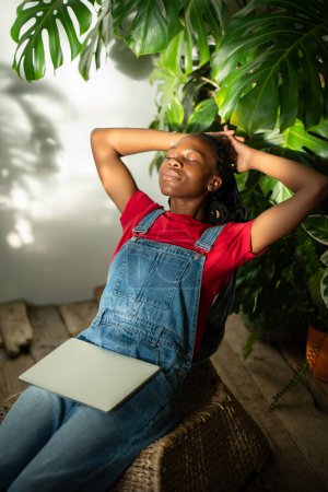 Entspannte afrikanisch-amerikanische Frau mit geschlossenen Augen und Händen hinter dem Kopf nach der Fernarbeit zu Hause. Zufriedene schwarze Freelancerin macht Pause auf Stuhl mit Laptop auf Knien im urbanen Dschungel-Interieur.