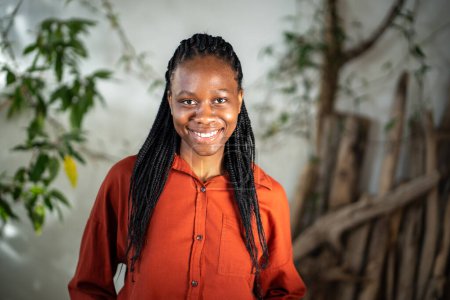 Cheerful African American young woman standing alone in room with green plants looking at camera, having wide white toothy charming smile, hair in braids, poses indoors. Happy portrait female.