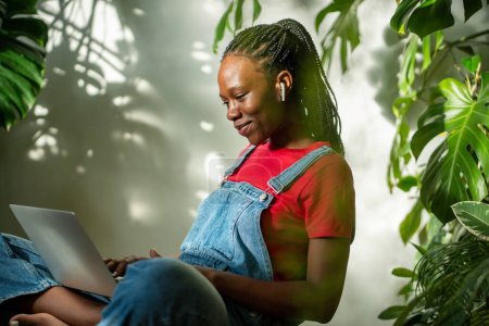 Foto de Mujer afroamericana agradable con el ordenador portátil en las piernas cruzadas sonriendo mirar la pantalla viendo vídeo en los auriculares se sienta en la habitación con las plantas. Chica negra satisfecha desplazándose por las redes sociales de Internet en la computadora - Imagen libre de derechos