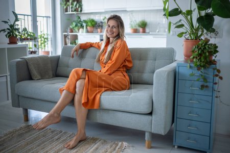 Portrait glückliche skandinavische Frau auf dem Sofa sitzend und lächelnd in die Kamera blickend, entspannt die Zeit genießend. Zufriedene Frauen entspannen sich auf der Couch im Wohnzimmer. Pflanzenliebhaber, gemütliches Zuhause, psychische Gesundheit.