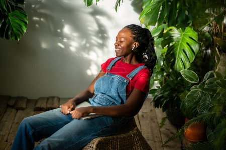 Une femme afro-américaine joyeuse porte un casque, se repose, sourit et s'assoit dans la chambre avec des plantes d'intérieur, tient un ordinateur portable sur les tours. Femme noire se détendre, écouter de la musique, prendre une pause du travail à la maison jardin