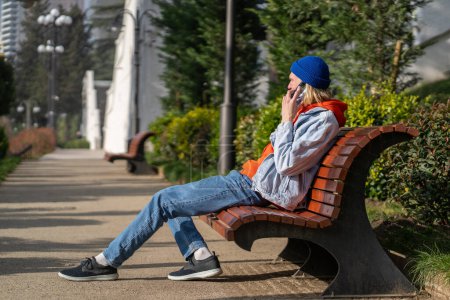 Téléchargez les photos : Jeune homme détendu faire conversation d'appel assis sur un banc en bois portant un bonnet et des vêtements en denim dans le parc de la ville. scandinave hipster guy parler bavarder sur téléphone mobile repos entourent par des plantes urbaines - en image libre de droit