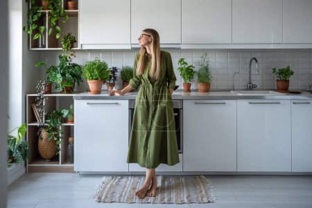 Photo for Pensive middle aged woman thinking looking at window, enjoys the tranquillity of home. Thoughtful female in glasses relaxed stand in cozy kitchen holding glass of water. Cozy interior with houseplant. - Royalty Free Image