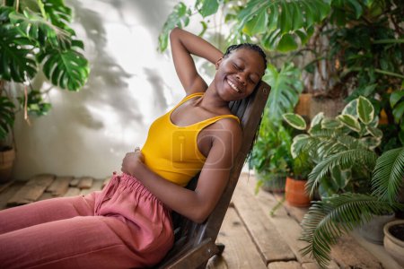 Joyful African American woman with smile resting sits on armchair in home garden. Pleased black female relaxing in greenhouse with houseplants. Slow life, mindfulness, stress free, plant lover concept