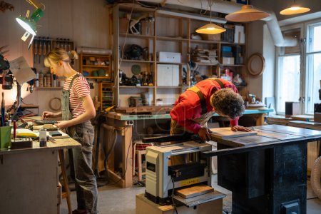 Family carpentry workshop, couple of carpenters working on wood in joinery. Female business owner calculating new order on calculator, focused man is busy doing woodwork. Woodworking industry. 
