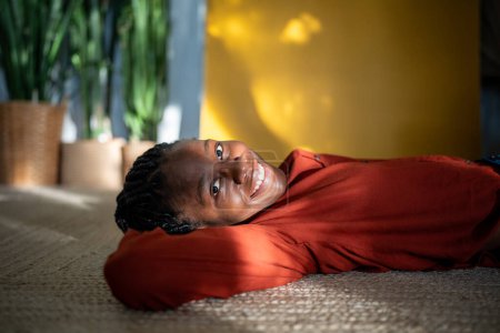 Carefree relaxed african american woman rest on carpet looking at camera smiling. Portrait serene pleased black young female lying on floor with hand under head feel happiness, enjoy sunny day at home