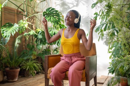 African American girl enjoying music with headphones dances while sitting on armchair in cozy room with urban jungle interior. Happy black female resting in home greenhouse listening favourite music.