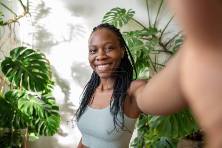 Joyful African American blogger girl make selfie on smartphone while standing in sunny room with houseplants. Contented black vlogger woman takes pictures of herself with wide smile on mobile phone