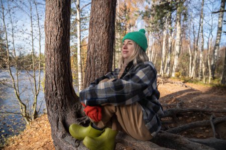 Foto de Mujer de mediana edad contemplando disfrutar de la naturaleza en el parque sentado en las raíces de los árboles. Mujer relajada en ropa de abrigo descansando en el bosque de otoño en la orilla del lago. Vida lenta, concepto de retiro - Imagen libre de derechos