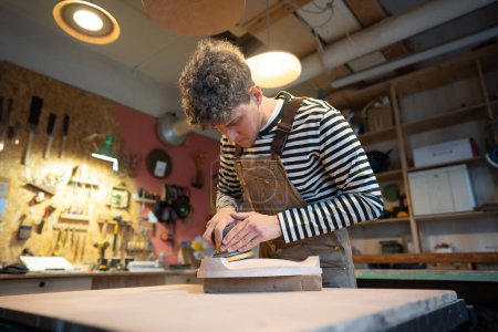 Holzschleifen mit Orbitalschleifer in der Werkstatt. Konzentrierter Tischler poliert Holzsitz eines zukünftigen Stuhls mit Elektroschleifer. Tischlerei. Möbelproduktion